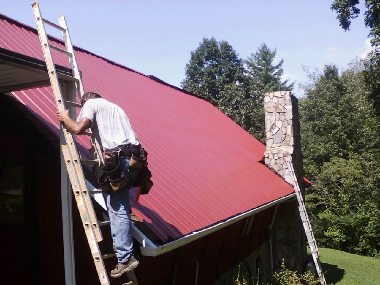Roofer on a ladder beside a red metal roof and stone chimney — Soffit & Fascia Repair by Roofing Brothers in La Salle